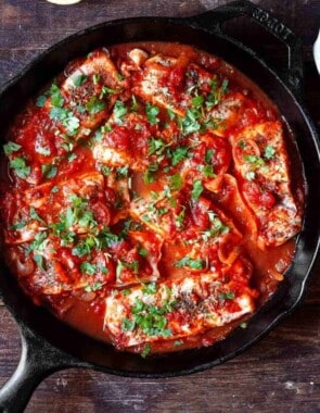 Greek fish with tomatoes and onions in a cast iron pan. parsley and lemon wedges to the side for serving