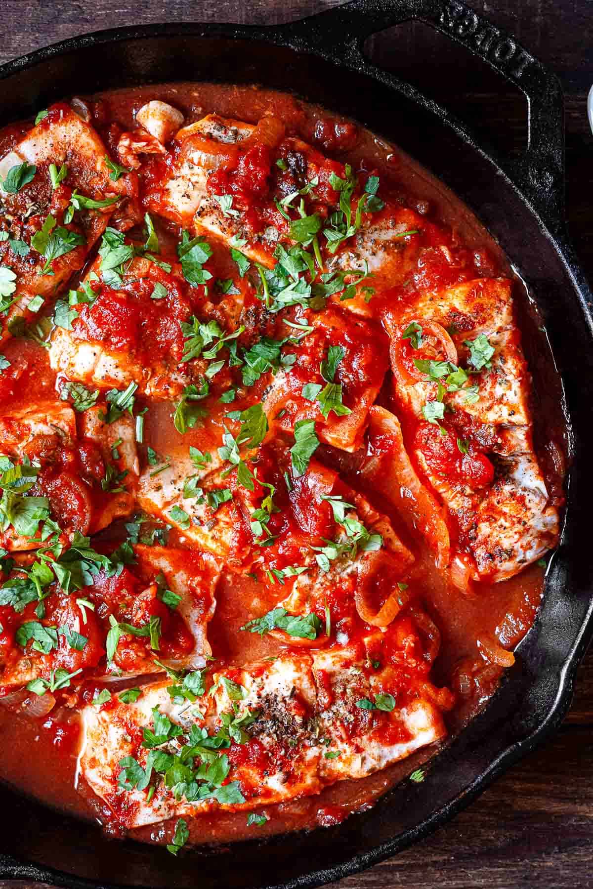 close up of 8 pieces of cooked white fish in a tomato sauce in a cast iron skillet.