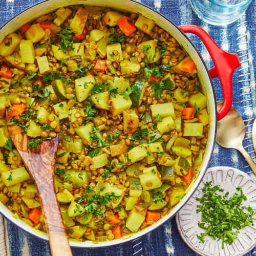 vegan lentil vegetable stew in a dutch oven with a wooden spoon, next to a bowl of chopped parsley, an empty bowl and two spoons.