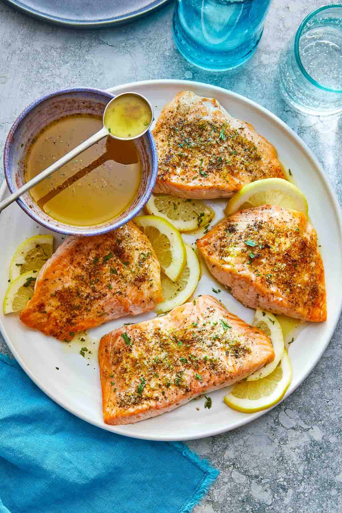 four broiled salmon fillets on a plate with lemon slices and a bowl of greek ladolemono dressing with a spoon..