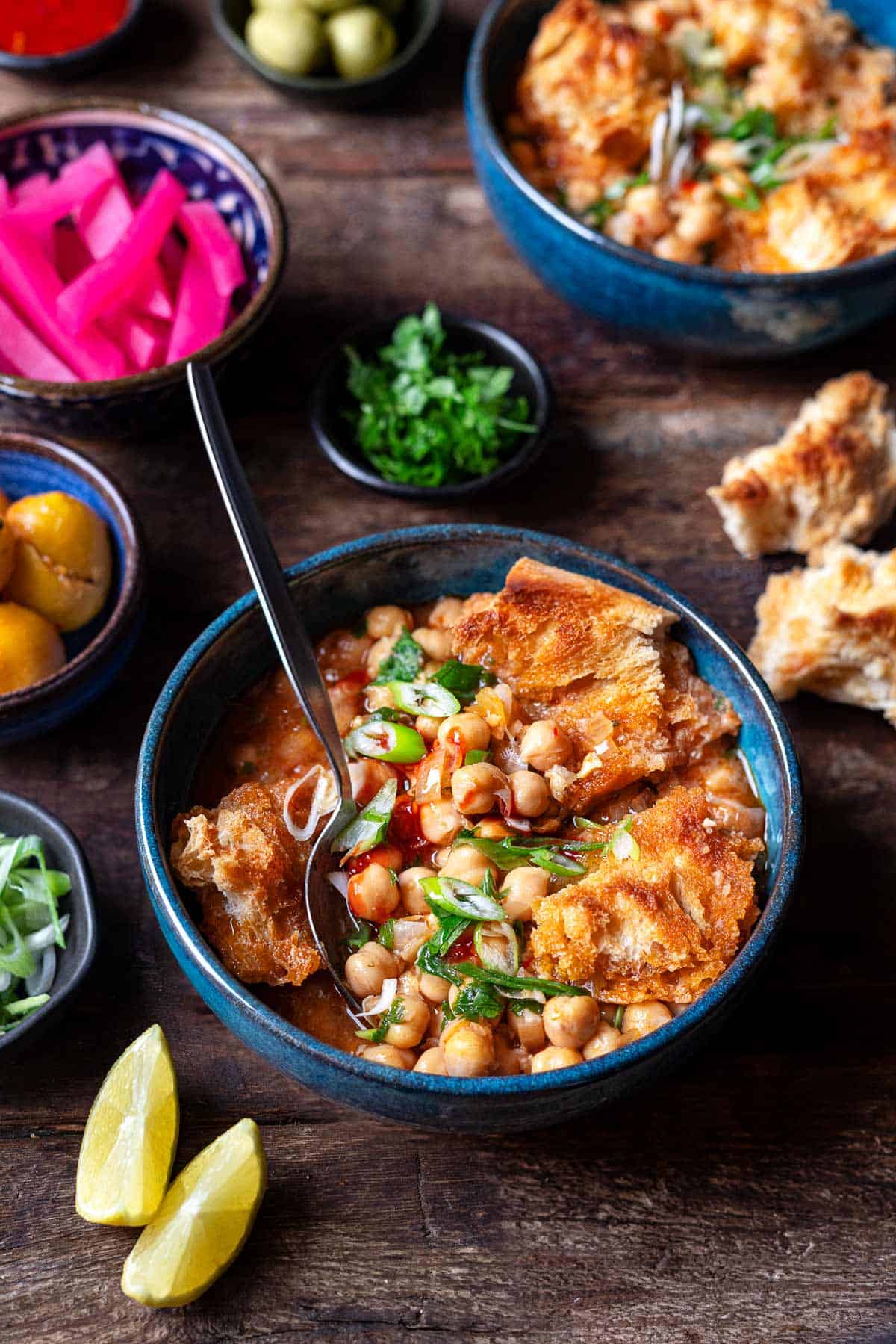 lablabi tunisian chickpea stew in a bowl, next to bowls of chopped green onions, preserved lemons, pickled turnips and pieces of torn up toasted bread.