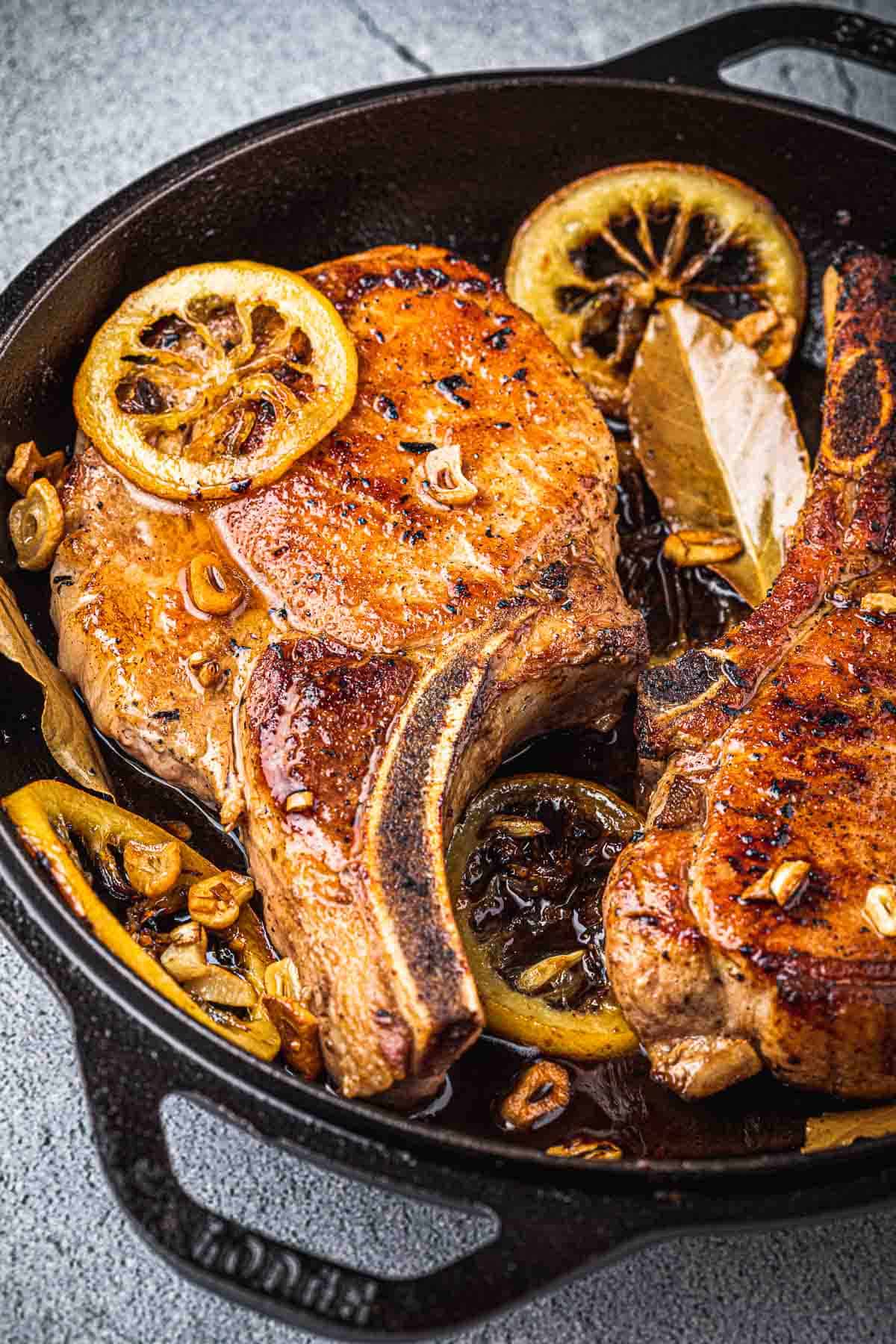 close up of two pan seared pork chops in a cast iron skillet with bay leaves and fried lemon rings.