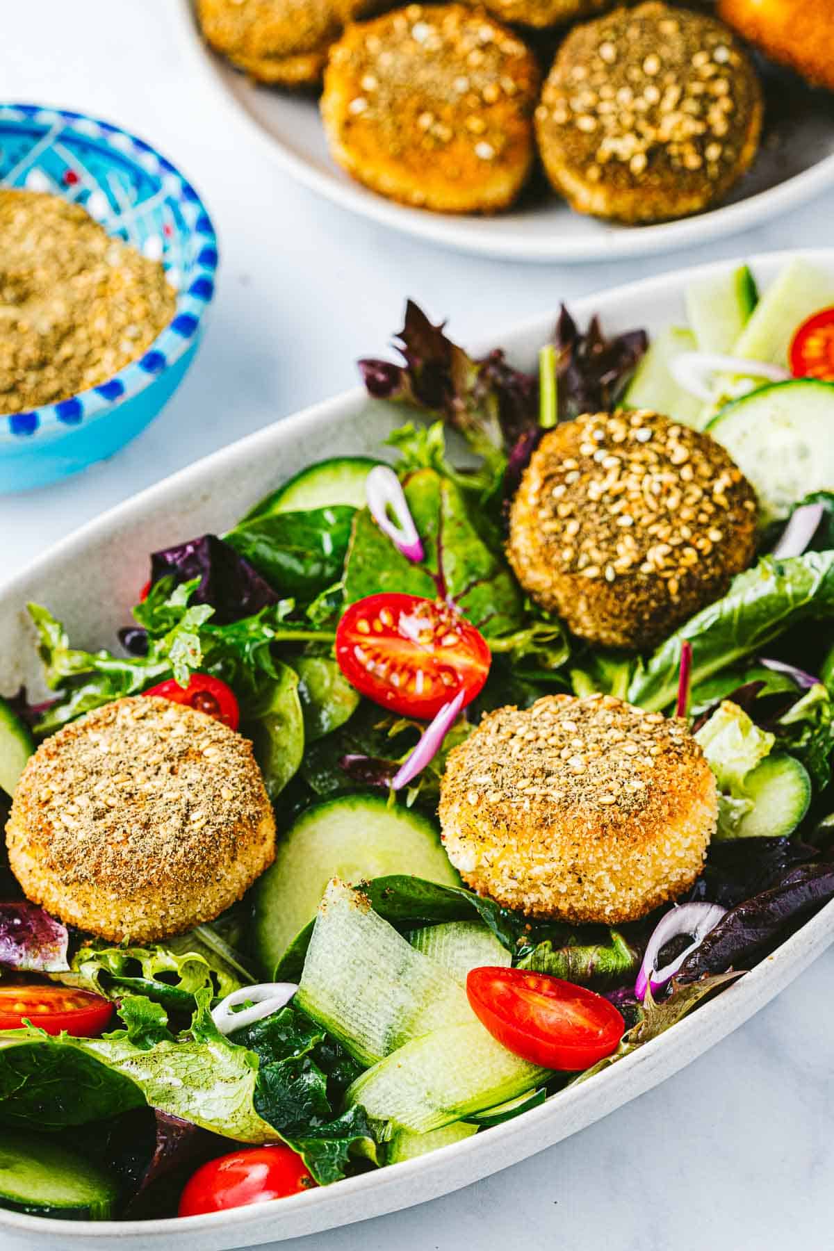 3 whole fried goat cheese slices on a bed of salad in a bowl next to a bowl of za'atar and a plate of fried goat cheese slices.