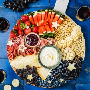 overhead photo of a complete fruit charcuterie board surrounded by 3 glasses of wine, grapes, a bowl of strawberries, and a group of crackers.