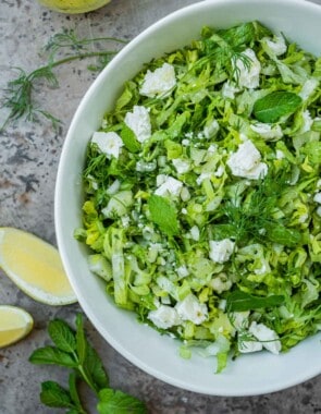 overhead photo of maroulosalata in a white serving bowl, next to a jar of dressing, sliced lemons, dill and mint.