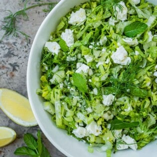 overhead photo of maroulosalata in a white serving bowl, next to a jar of dressing, sliced lemons, dill and mint.