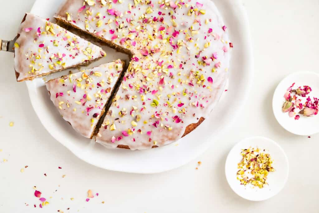 Overhead shot of Persian love cake on a platter with two slices cut out and rose petals and pistachios on the side.