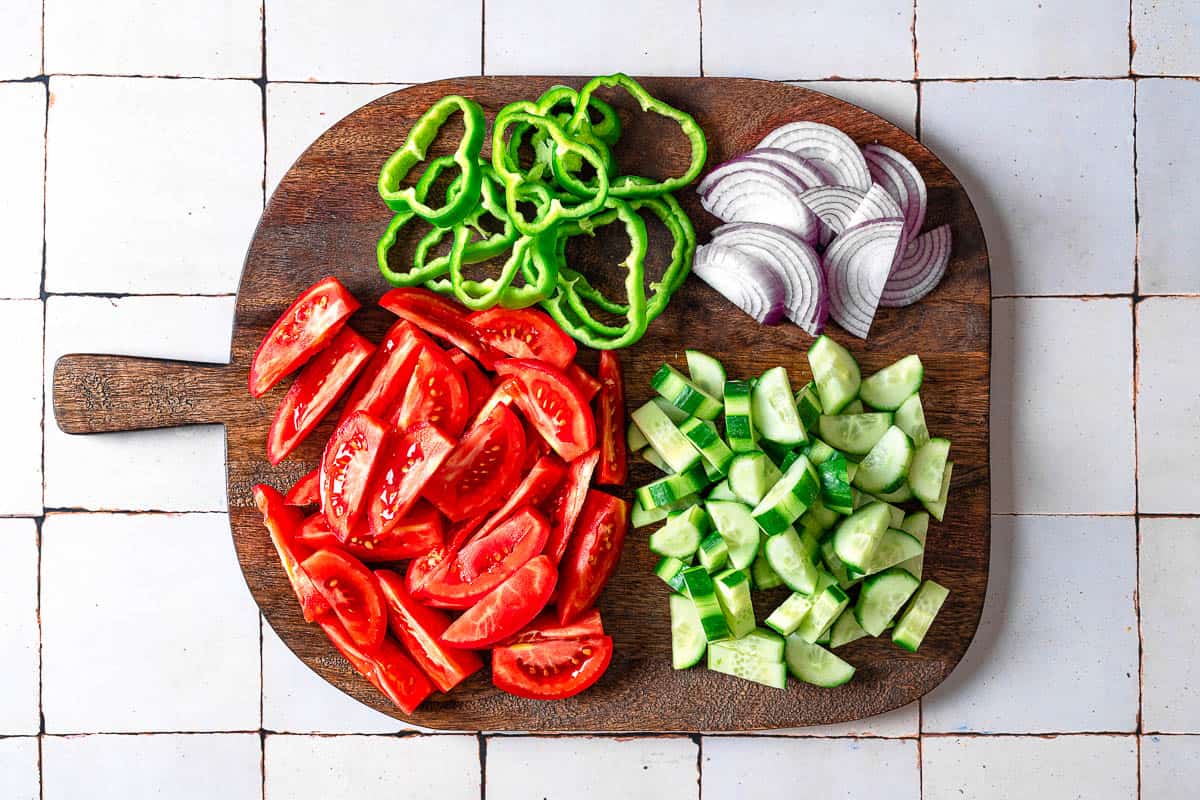 sliced green pepper, tomatoes, cucumbers and red onions on a wooden cutting board.