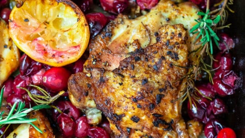 a close up of baked cranberry chicken topped with fresh cranberries, sprigs of fresh rosemary and lemon slices.