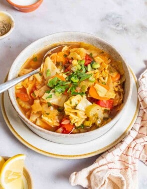 detox cabbage soup in a bowl with a spoon surrounded by small bowls of salt, pepper and lemon wedges and a cloth napkin.