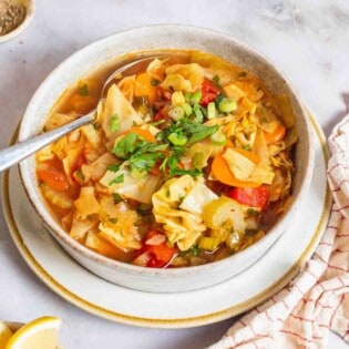 detox cabbage soup in a bowl with a spoon surrounded by small bowls of salt, pepper and lemon wedges and a cloth napkin.