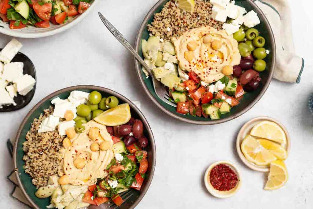 An overhead photo of 2 mediterranean bowls with quinoa with forks in them. These are surrounded by bowls of lemon slices, aleppo pepper, feta cheese and a Mediterranean cucumber tomato salad.