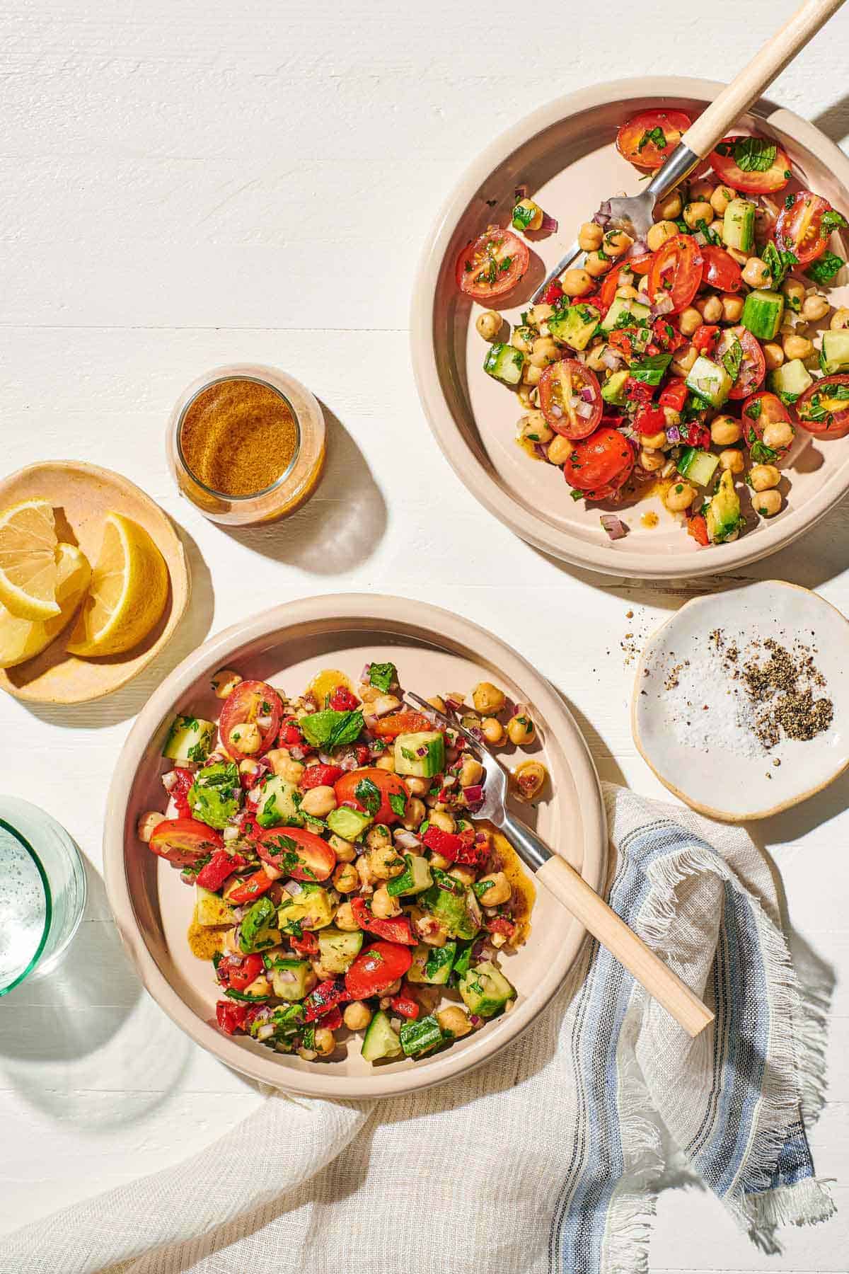 an overhead photo of two bowls of vegan chickpea salad with forks next to a small plate of salt and pepper, a small bowl of lemon wedges, a jar of dijon dressing, a glass of water and a cloth napkin.