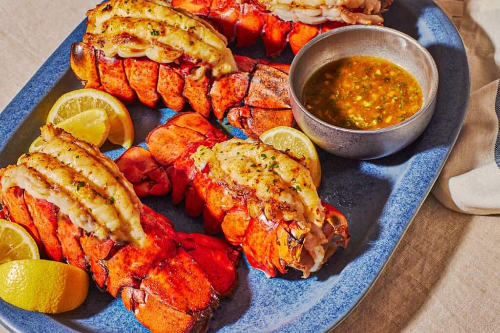 A close up of 4 broiled lobster tails on a blue serving platter lemon wedges and butter sauce in a small bowl.