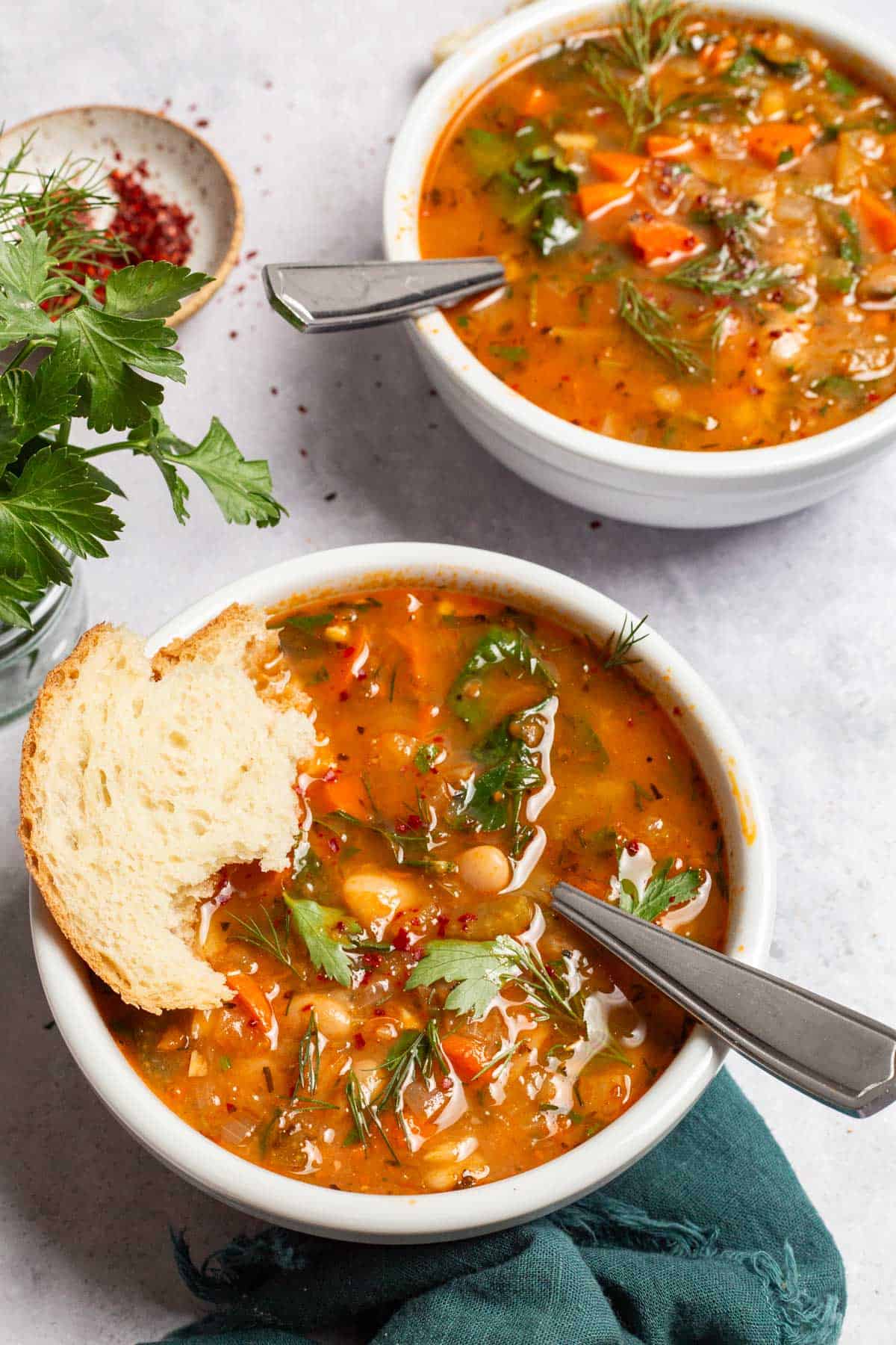 two bowls of Mediterranean white bean soup with spoons next to some parsley and a small bowl of aleppo pepper. One bowl of soup also has a piece of crusty bread in it.