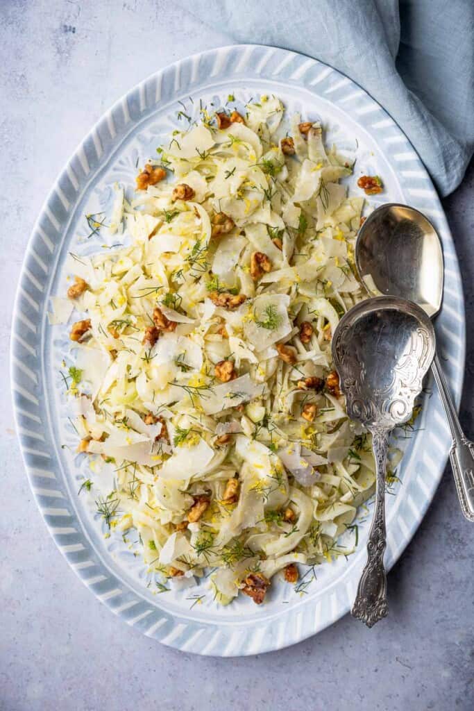 Overhead shot of fennel salad on a blue and white serving platter with two silver serving spoons.