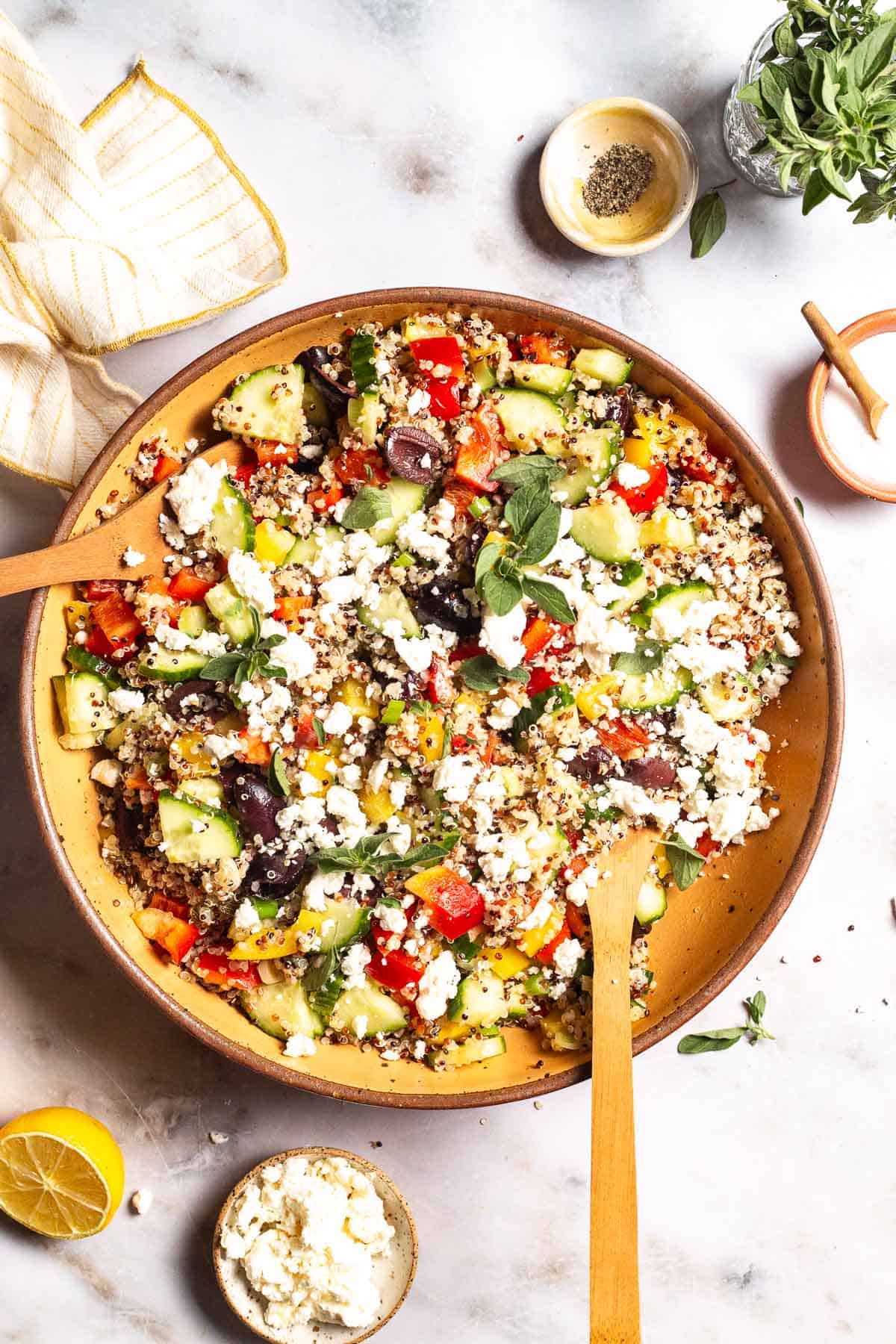 An overhead photo of quinoa salad in a large bowl with wooden serving utensils. This is surrounded by small bowls of feta cheese, salt and pepper as well as a lemon half, a jar of fresh oregano, and a cloth napkin.