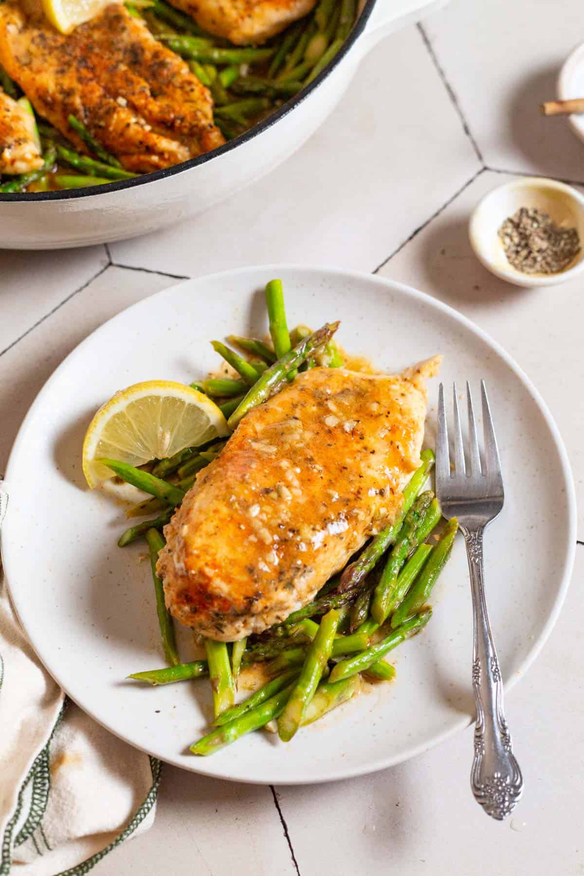 A photo of a serving of chicken and asparagus on a plates with a lemon wedge and a fork. Next to this are bowls of salt and pepper, a kitchen towel, and a skillet with the rest of the chicken and asparagus.