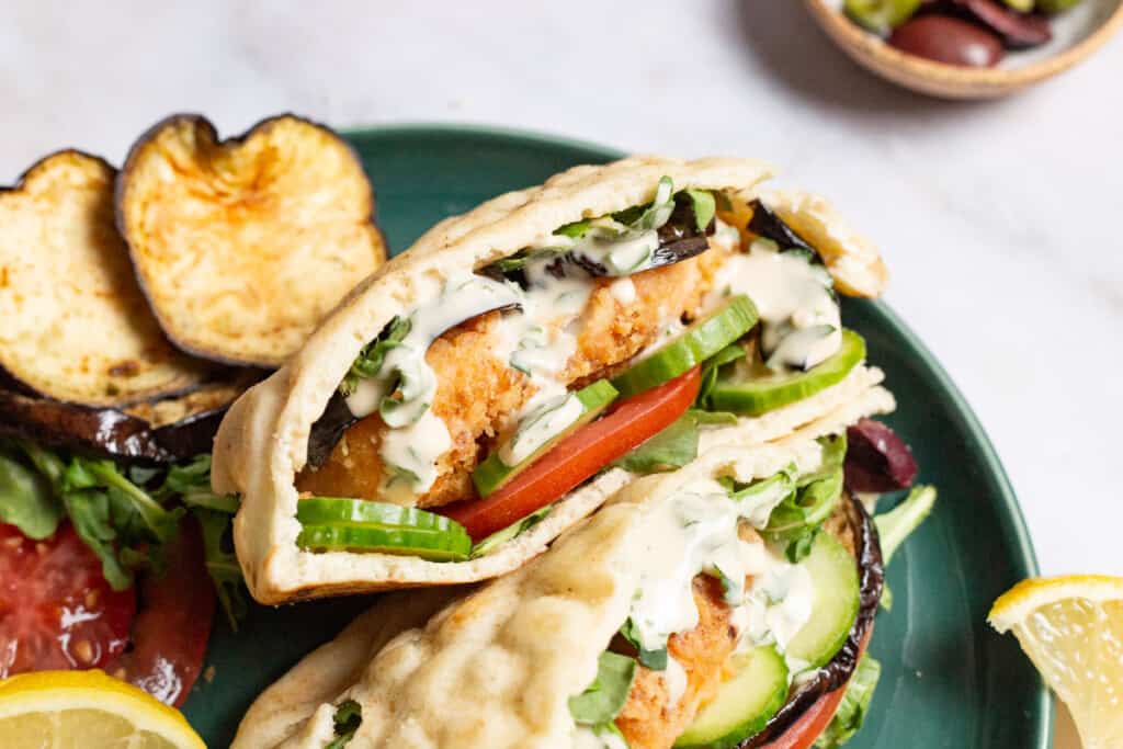 A close up of two fried fish sandwiches on a plate with sides of roasted eggplant slices, tomato, and a lemon wedge.