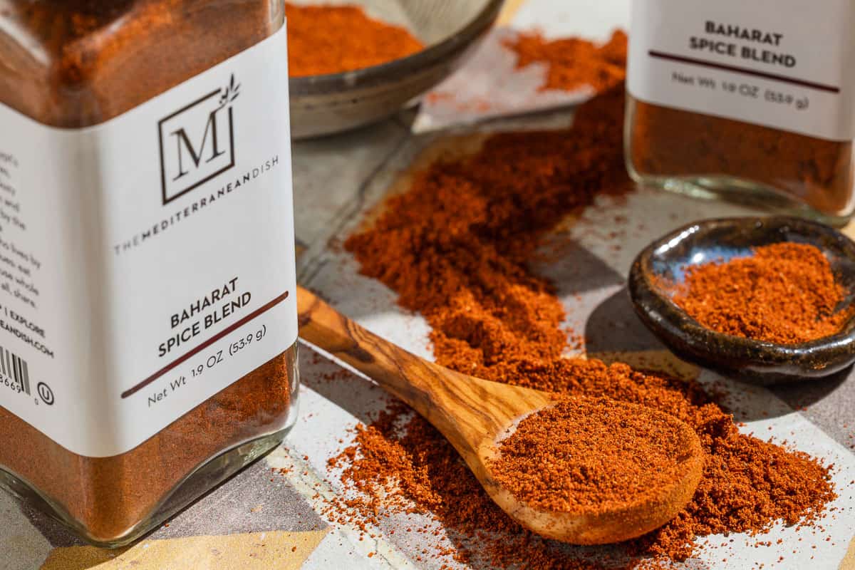 Baharat spice blend on a spoon and in a small bowl. Next to this are two jars of the spice from The Mediterranean Dish.