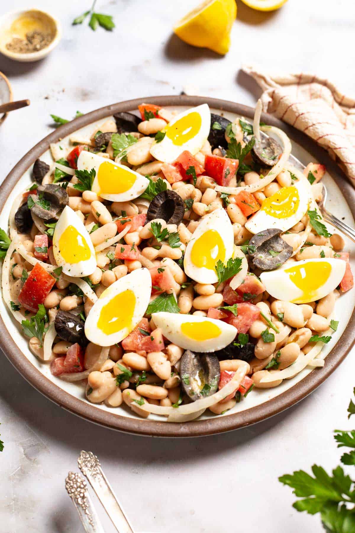 Cannellini bean salad on a serving platter with a fork. Next to this is a small bowl of pepper, 2 lemon halves, and a cloth napkin.