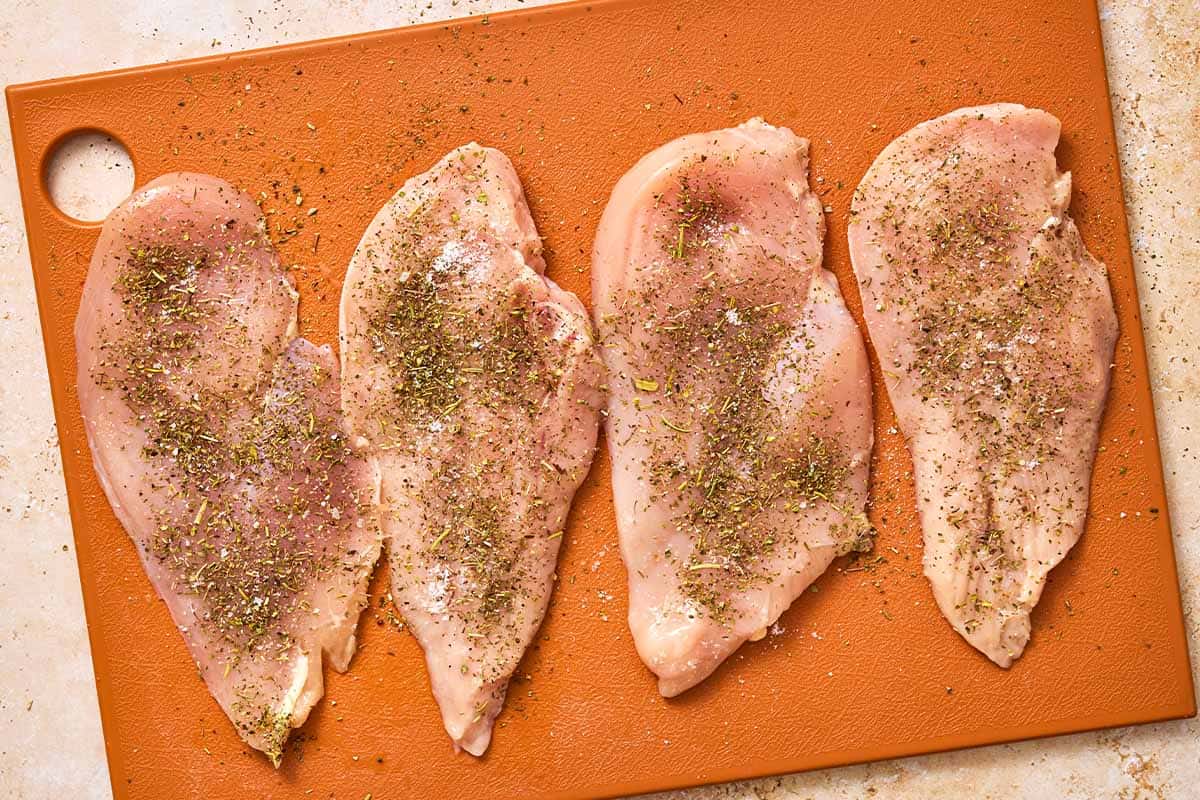 4 seasoned uncooked chicken cutlets on a cutting board.