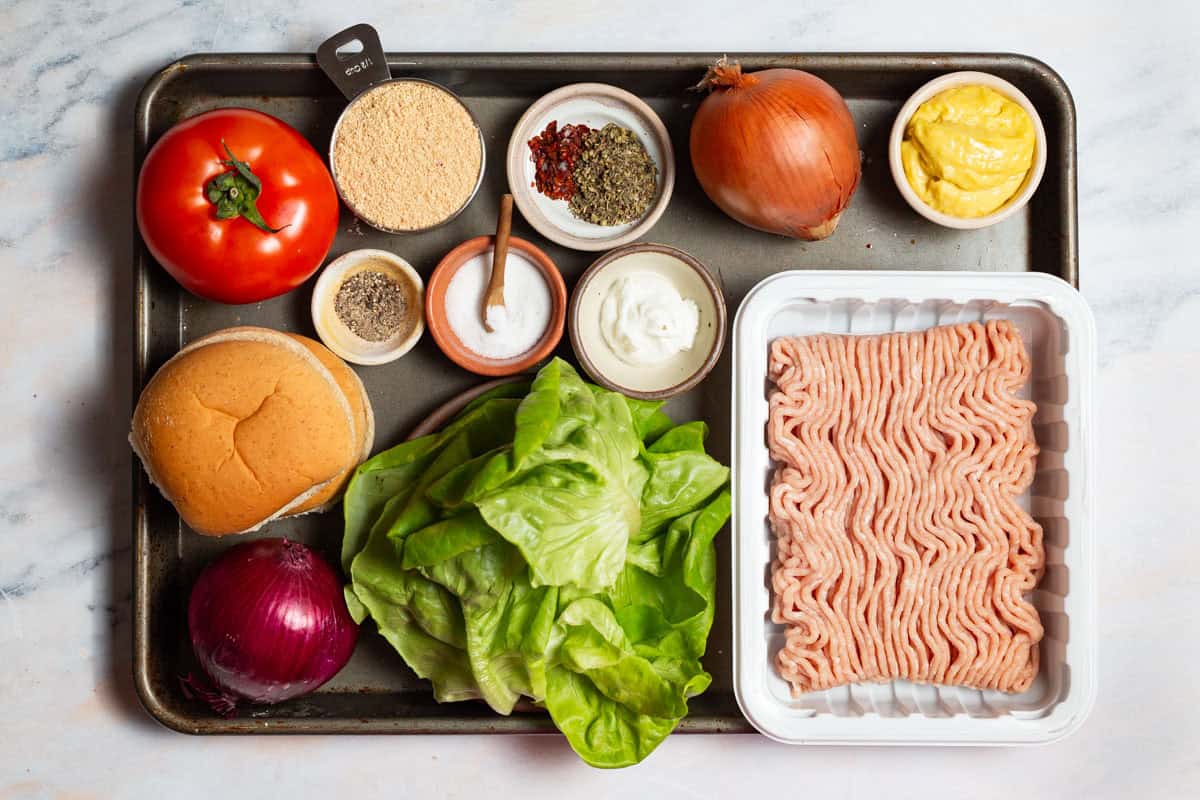 Ingredients for chicken burgers including ground chicken, bread crumbs, greek yogurt, yellow onion, italian seasoning, urfa biber salt, pepper, olive oil, buns, lettuce, a tomato and red onion.