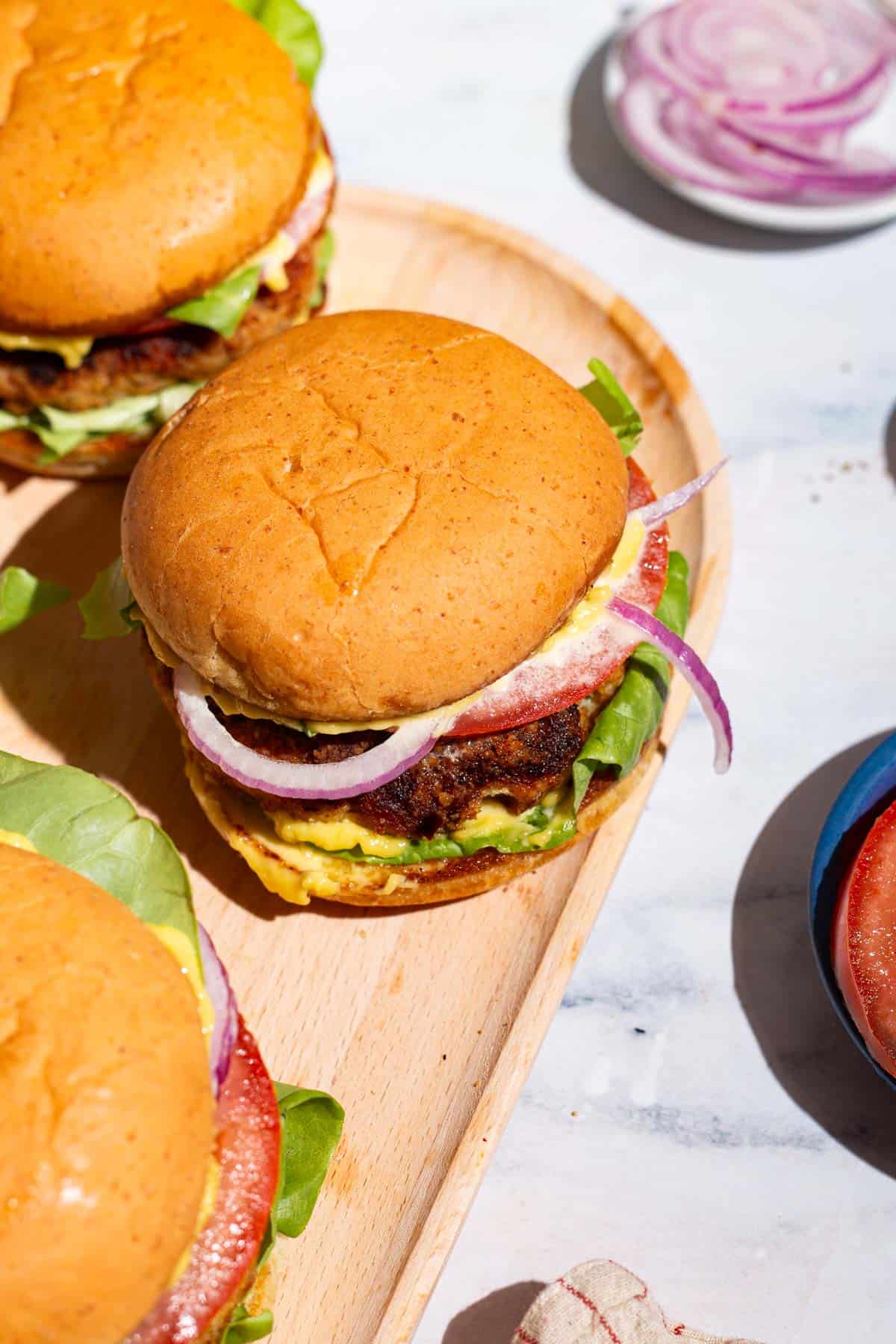 3 chicken burgers on a wooden serving platter. Next to this is a plate of sliced onions.