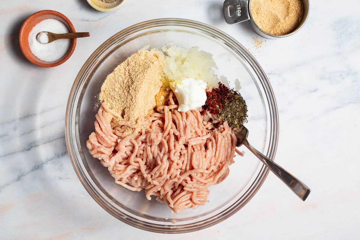 The ingredients for chicken burgers in a mixing bowl with a fork just before being combined. Next to this is a 1/2 cup measuring cup and bowls of salt and pepper.