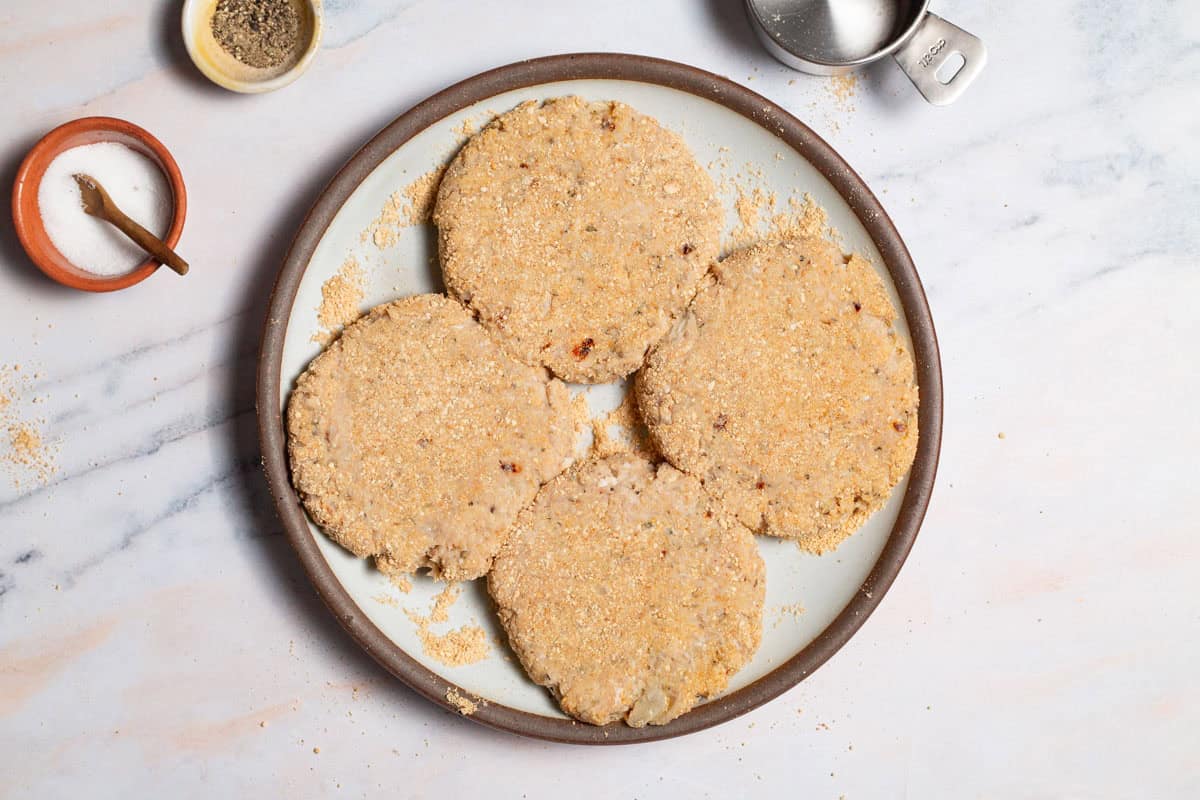 4 uncooked chicken burgers on a plate, surrounded by bowls of salt and pepper and an empty 1/2 cup measuring cup.