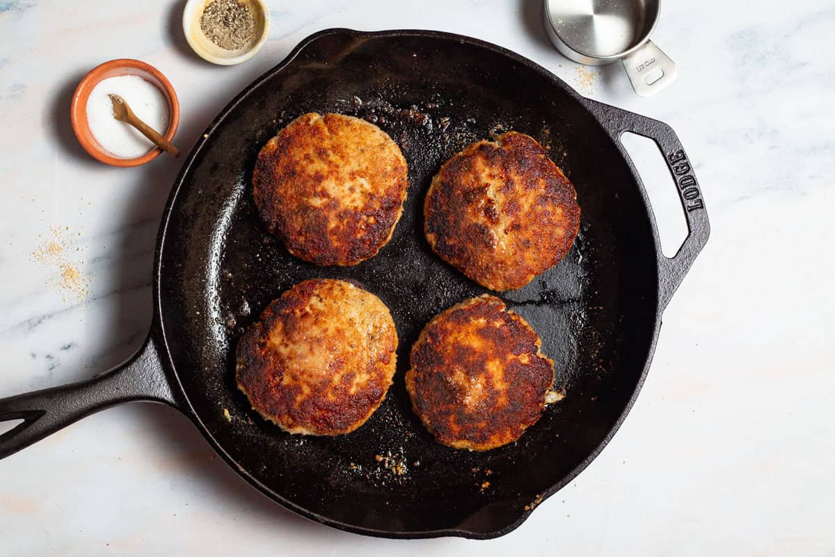 4 chicken burgers cooking in a cast iron skillet surrounded by bowls of salt and pepper and an empty 1/2 cup measuring cup.