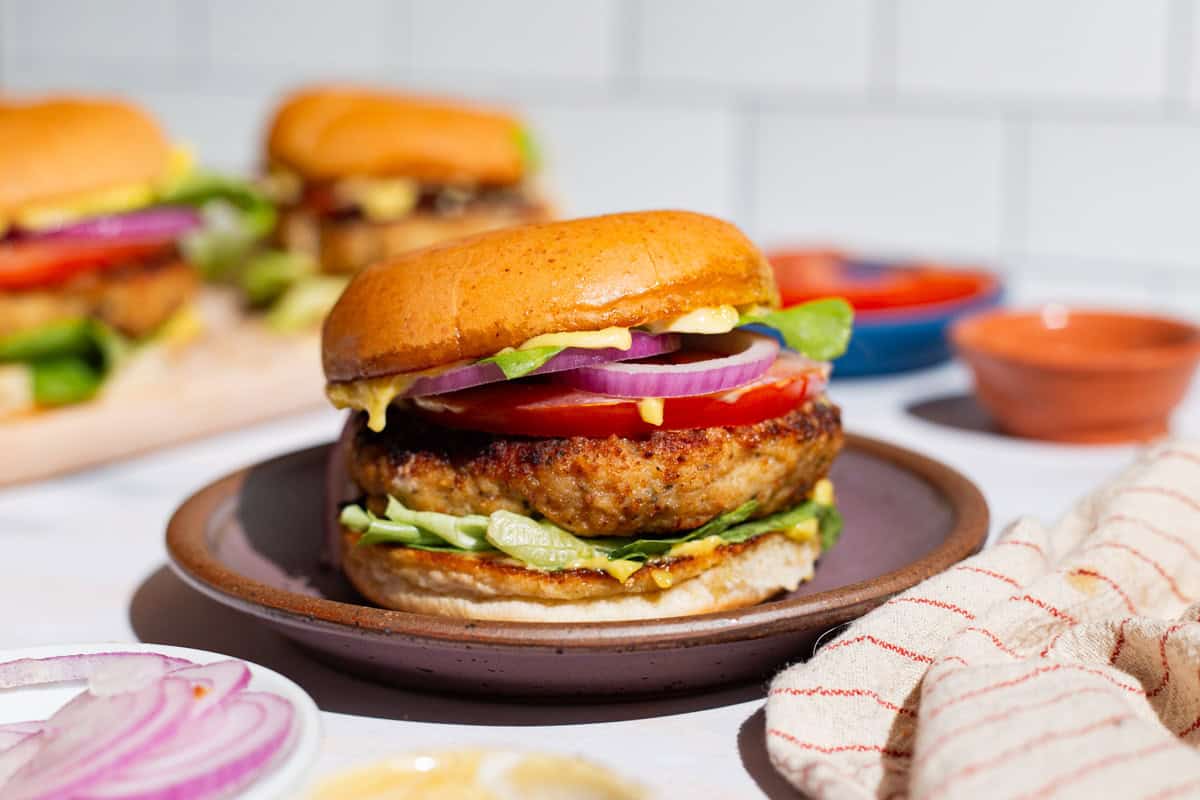 A close up of a chicken burger on plate next to a kitchen towel and a plate of sliced red onions.