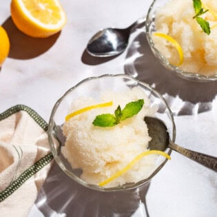 Two bowls of lemon sorbet, one with a spoon, and one next to a spoon. Next to these are 2 lemon wheels and a kitchen towel.