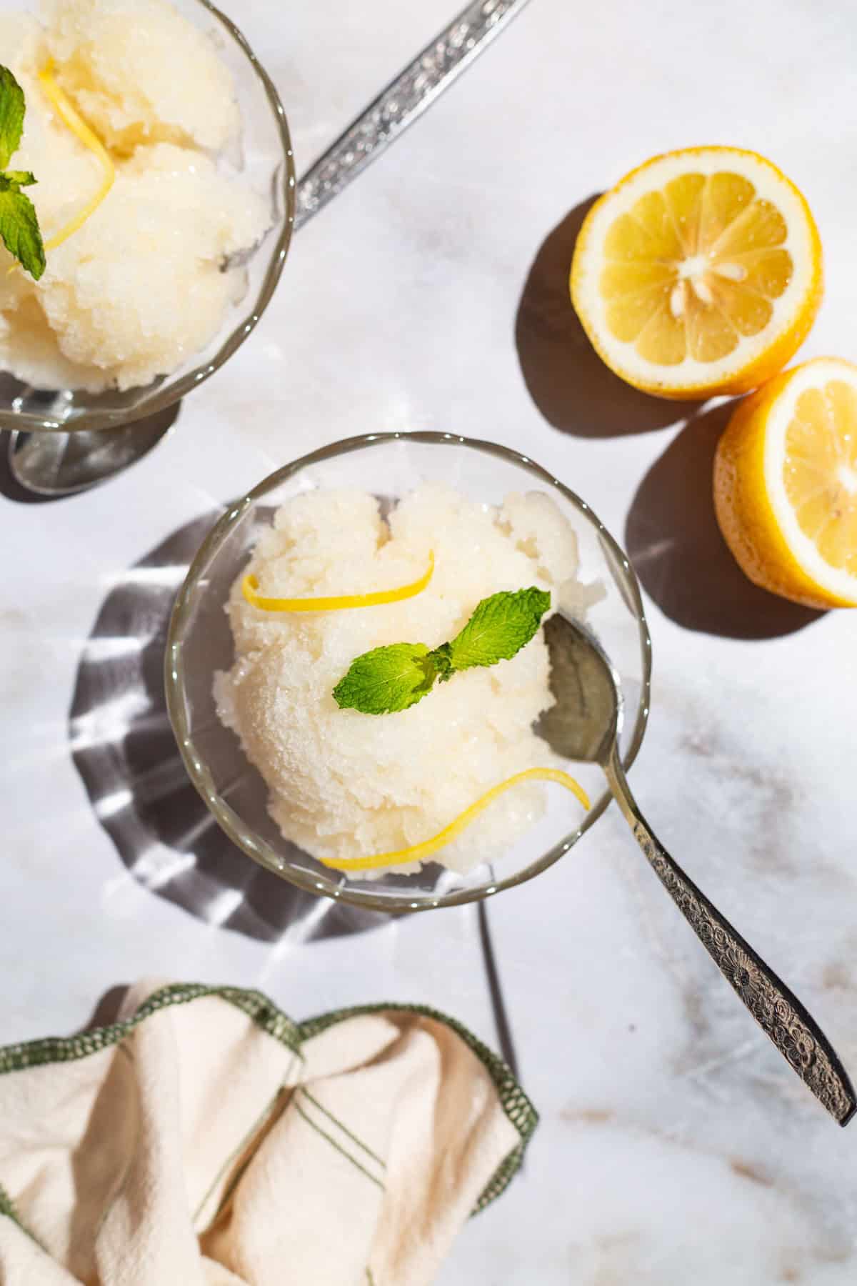 An overhead photo of 2 bowls of lemon sorbet, one with a spoon, and one next to a spoon. Next to these are 2 lemon wheels and a kitchen towel.