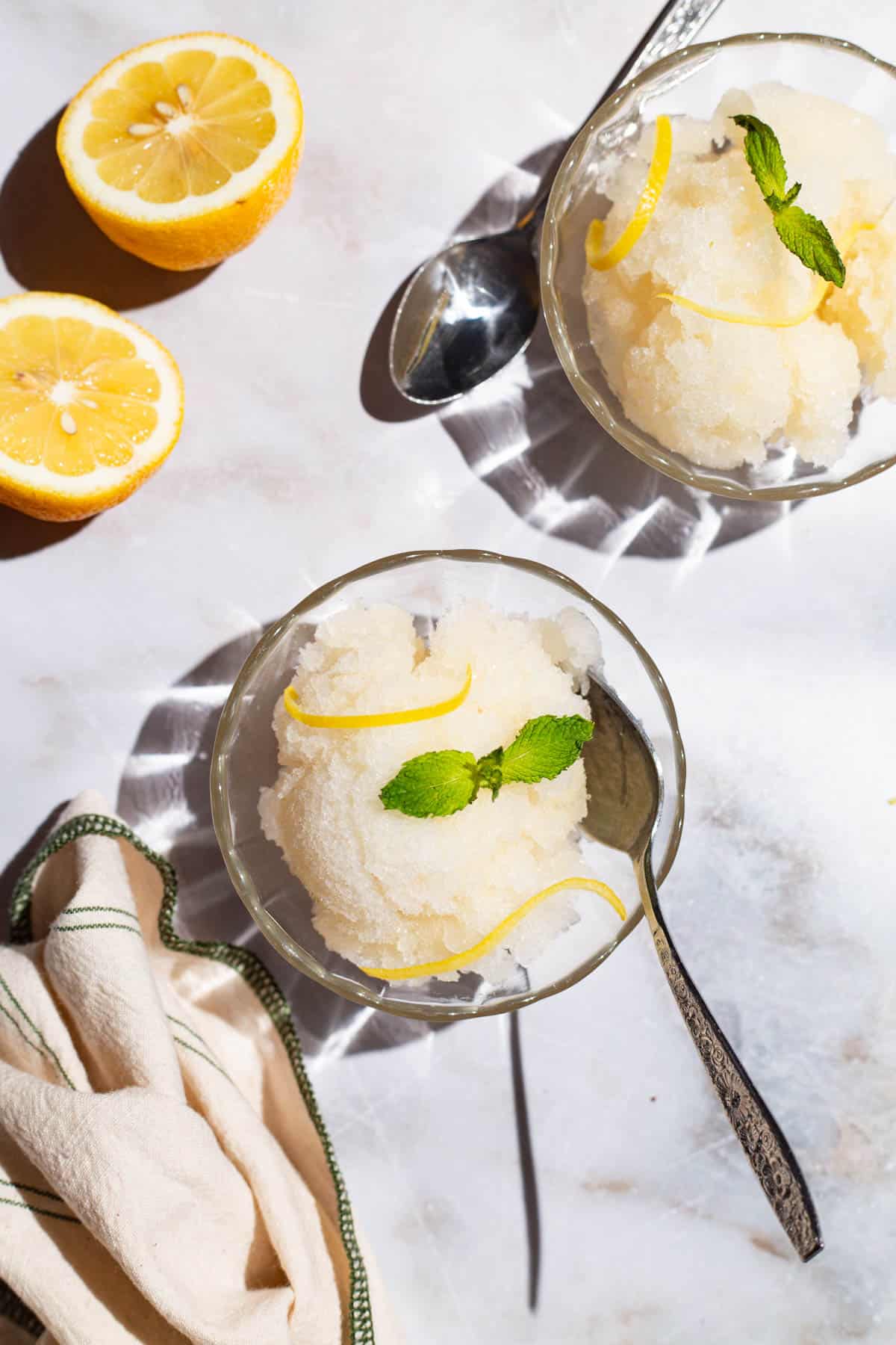 An overhead photo of 2 bowls of lemon sorbet, one with a spoon, and one next to a spoon. Next to these are 2 lemon wheels and a kitchen towel.
