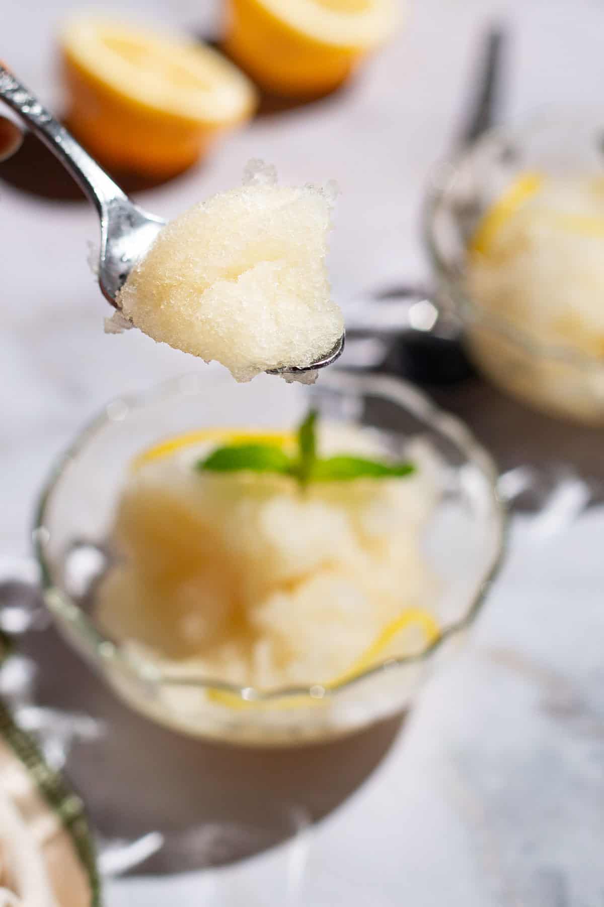A close up of a spoonful of lemon sorbet with two bowls of sorbet and 2 lemon halve in the background.