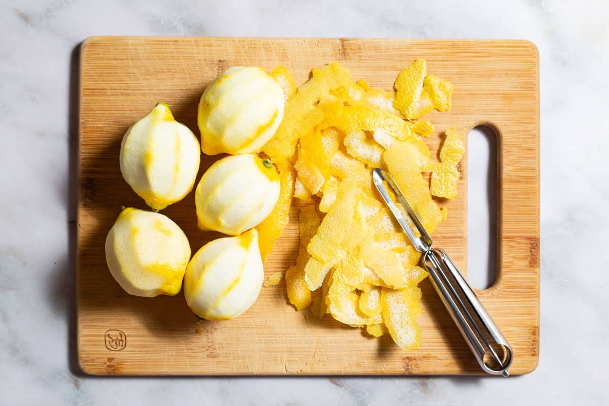 5 zested lemons next to a pile of lemon zest strips and a peeler on a wooden cutting board.