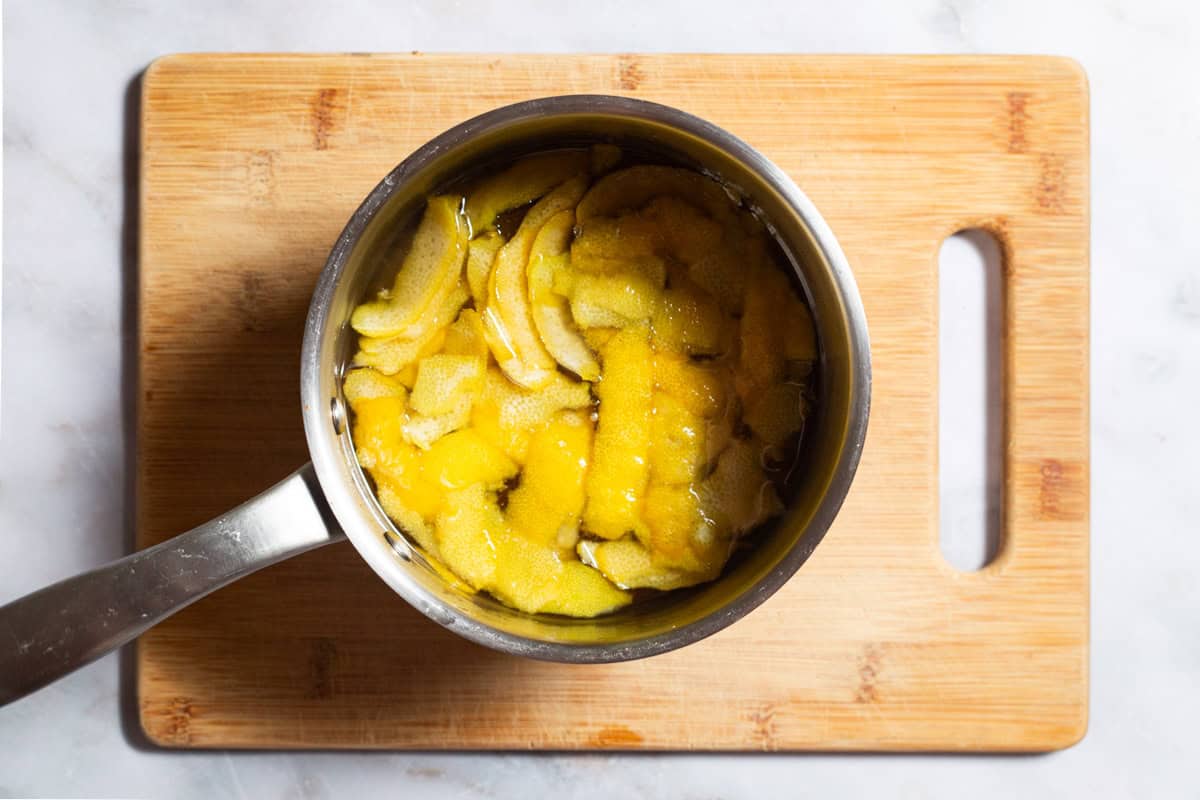 Water, sugar and lemon zest strips in a saucepan sitting on a wooden cutting board.
