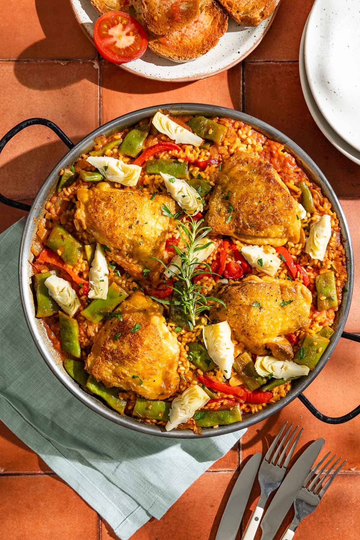 An overhead photo of chicken paella in a large paella pan. Next to this is a cloth napkin, knives and forks, a stack of 2 plates, and a plate with tomatoes and toasted bread.
