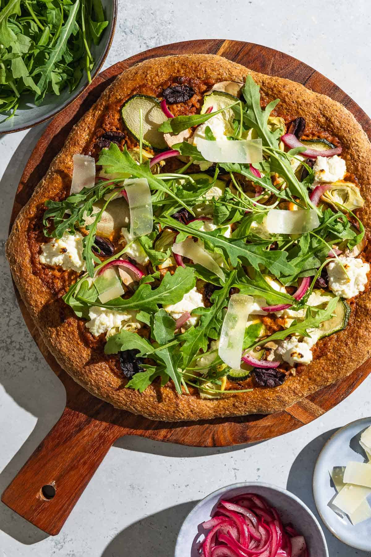 An overhead photo of a vegetarian pizza on a wooden serving platter next to bowls of arugula, pickled onions, and parmesan cheese.