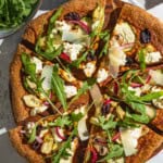 An overhead photo of a sliced vegetarian pizza on a wooden serving platter next to bowls of arugula and pickled onions.