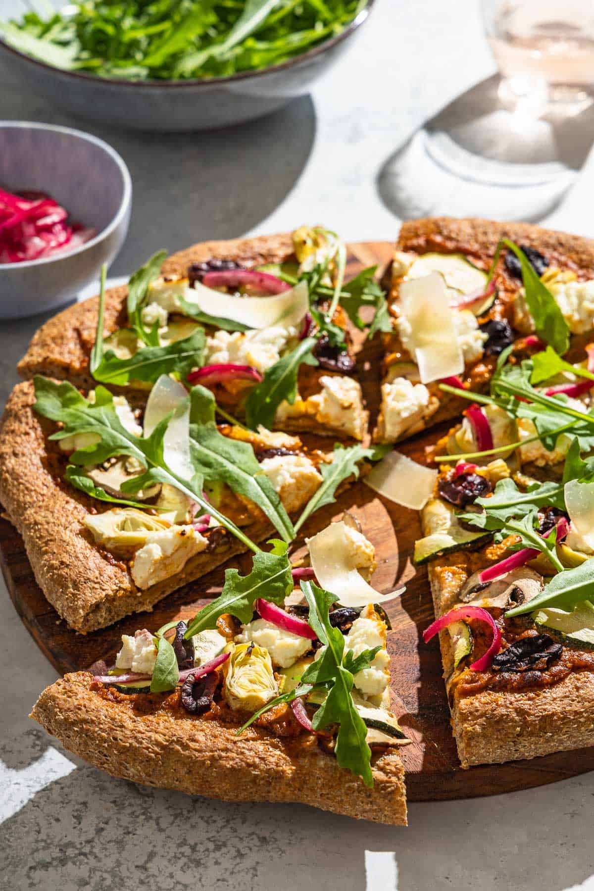 A close up of a sliced vegetarian pizza on a wooden serving platter next to bowls of arugula and pickled onions, and a glass of water.