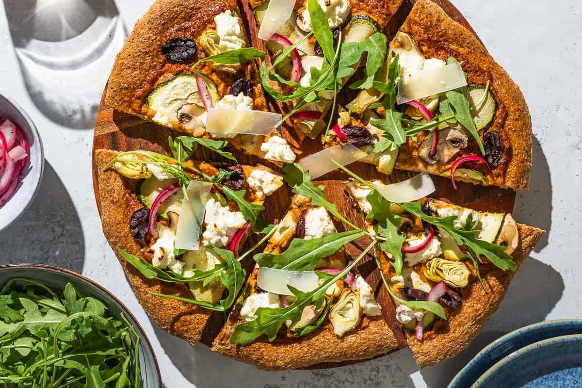 An overhead photo of a sliced vegetarian pizza on a wooden serving platter next to bowls of arugula and pickled onions.