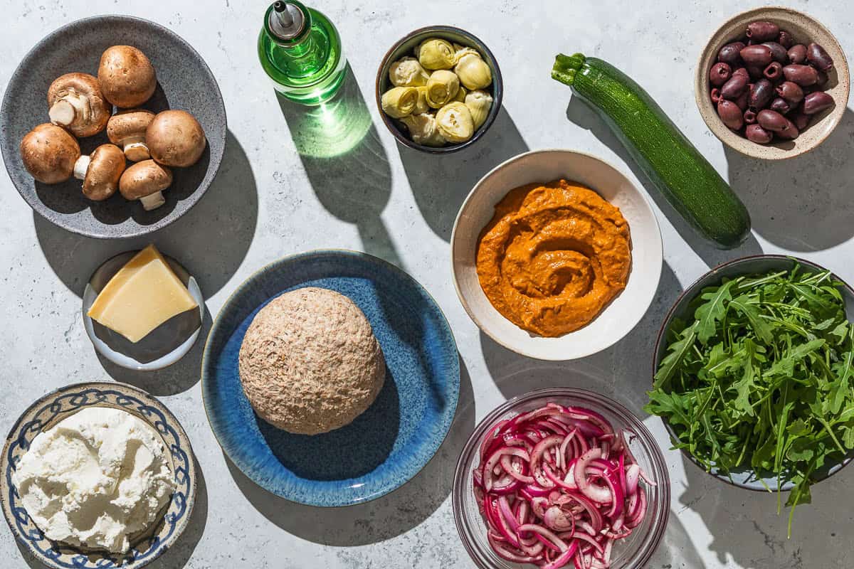 Ingredients for vegetarian pizza including whole wheat pizza dough, olive oil, romesco cause, zucchini, mushrooms, kalamata olives, artichoke hearts, ricotta cheese, pickled red onions, arugula, and parmesan cheese.