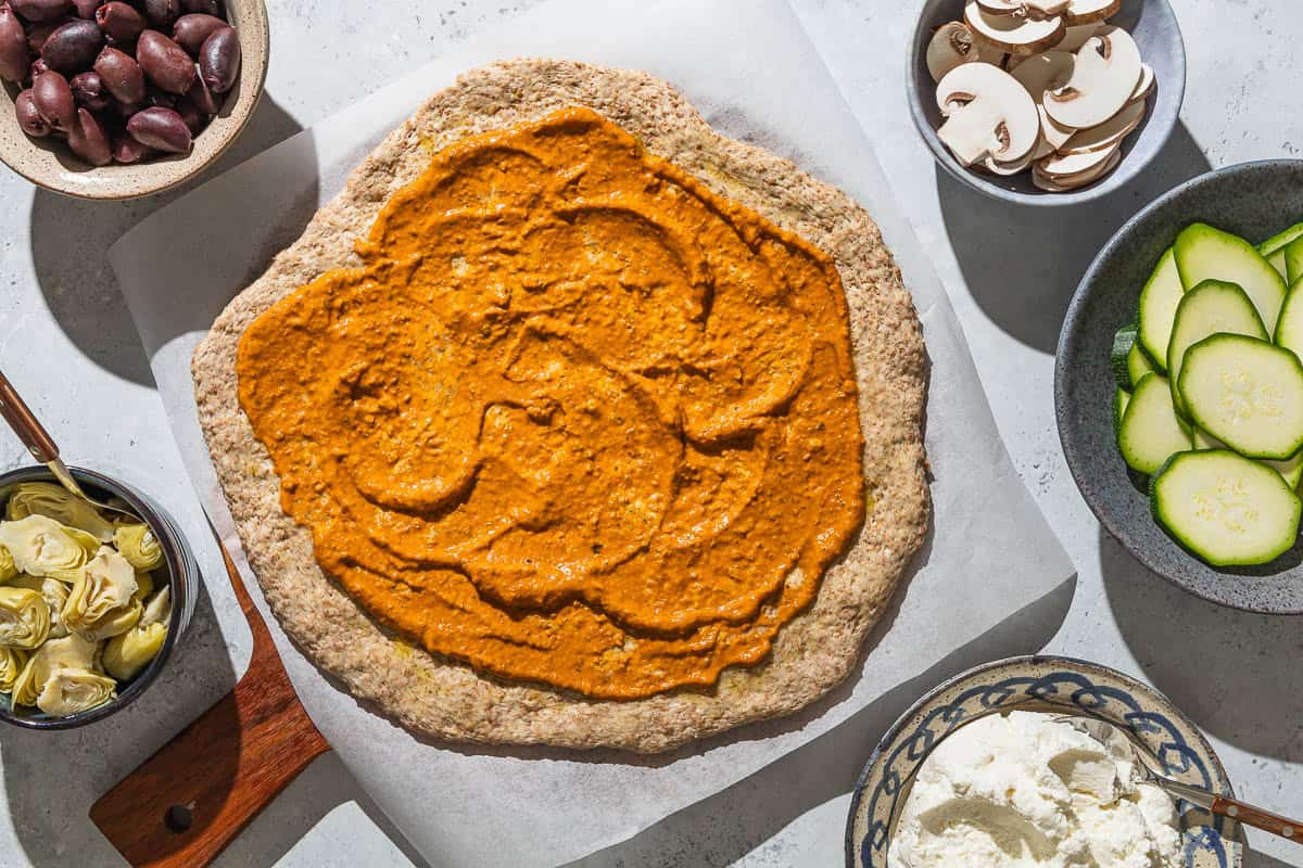 An overhead photo of an unbaked whole wheat pizza crust layered with romesco sauce on a parchment-lined wooden serving tray. Surrounding this are bowls of artichoke hearts, kalamata olives, mushrooms, zucchini and ricotta cheese.