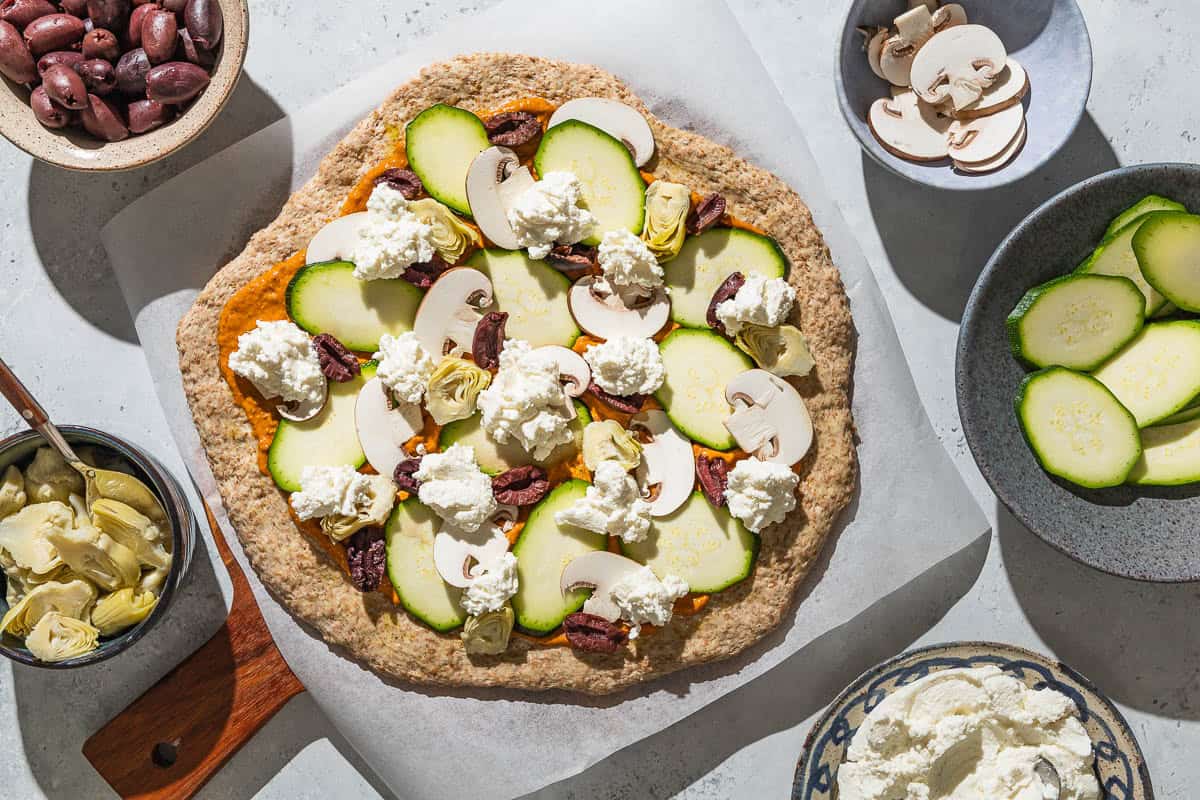 An overhead photo of an unbaked vegetarian pizza on a parchment-lined wooden serving tray. Surrounding this are bowls of artichoke hearts, kalamata olives, mushrooms, zucchini and ricotta cheese.