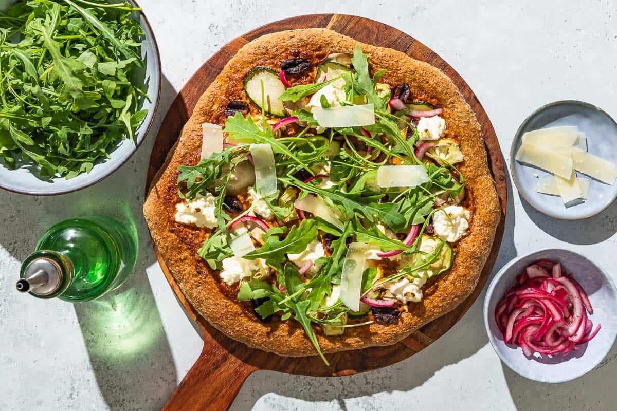 An overhead photo of a vegetarian pizza on a wooden serving platter next to bowls of arugula, pickled onions and parmesan cheese, and a bottle of olive oil.