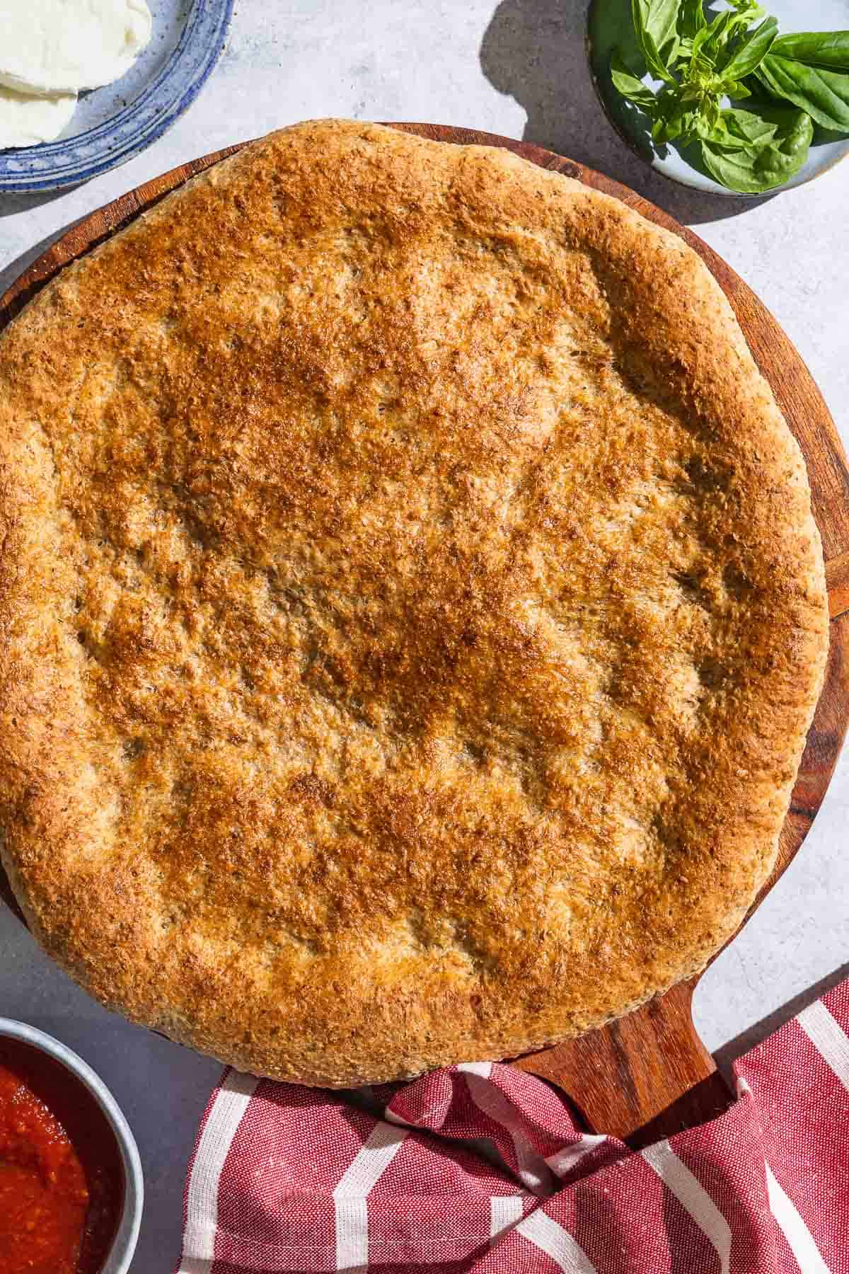 An overhead photo of baked whole wheat pizza crust on a wooden serving platter with a kitchen towel. Next to this is a bowl of pizza sauce, and plates with sliced mozzarella and basil.