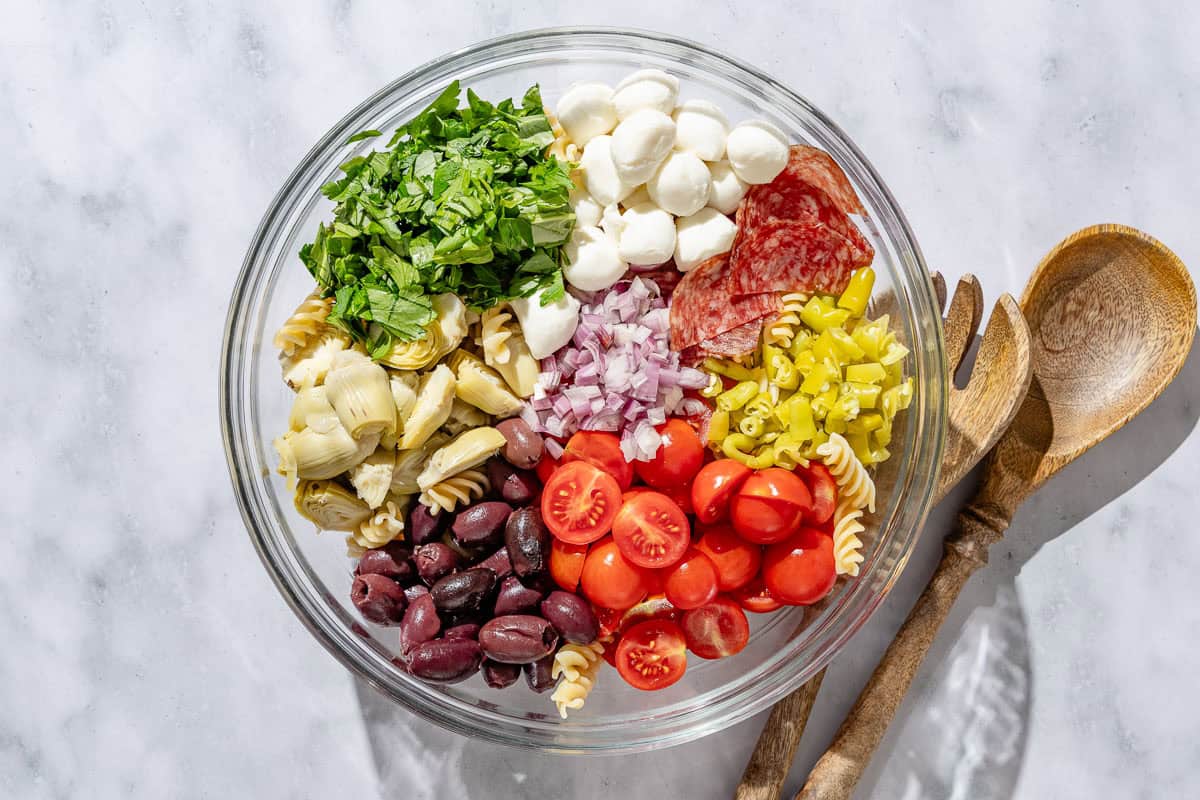 Mixing bowl with groups of mozzarella, parsley, basil, pepperoncini, cherry tomatoes, artichoke hearts, kalamata olives, and shallot before being mixed.