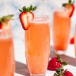 A close up of a strawberry bellini in a glass garnished with a strawberry slice surrounded by more glasses of the bellini and whole strawberries.